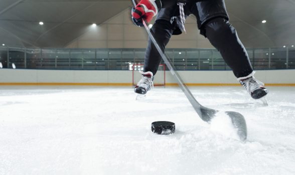 Male hockey player in sports uniform, gloves and skates moving down rink in front of camera against stadium environment while going to shoot puck